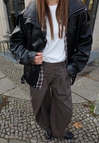 Chaqueta de cuero negra sobredimensionada sobre una camiseta blanca, combinada con pantalones marrones de pierna ancha que muestran un patrón de cuadros asomándose desde el interior.