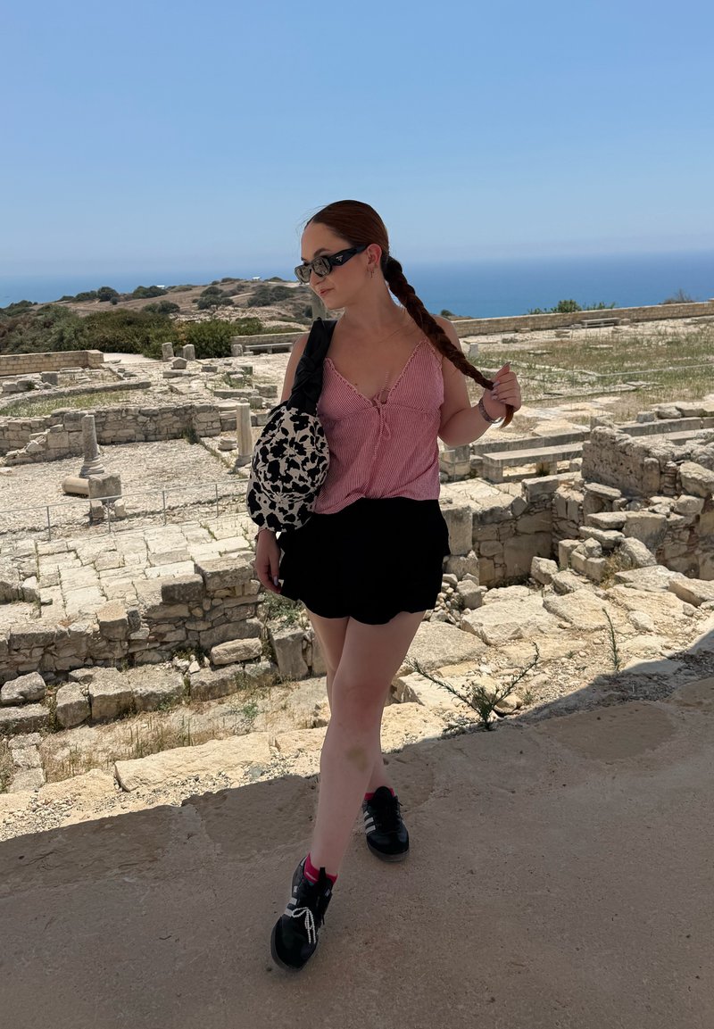Γυναίκα με μακριά πλεξούδα και γυαλιά ηλίου στέκεται ανάμεσα σε αρχαία πέτρινα ερείπια με θάλασσα και λόφους στο φόντο σε μια καθαρή ηλιόλουστη μέρα.
