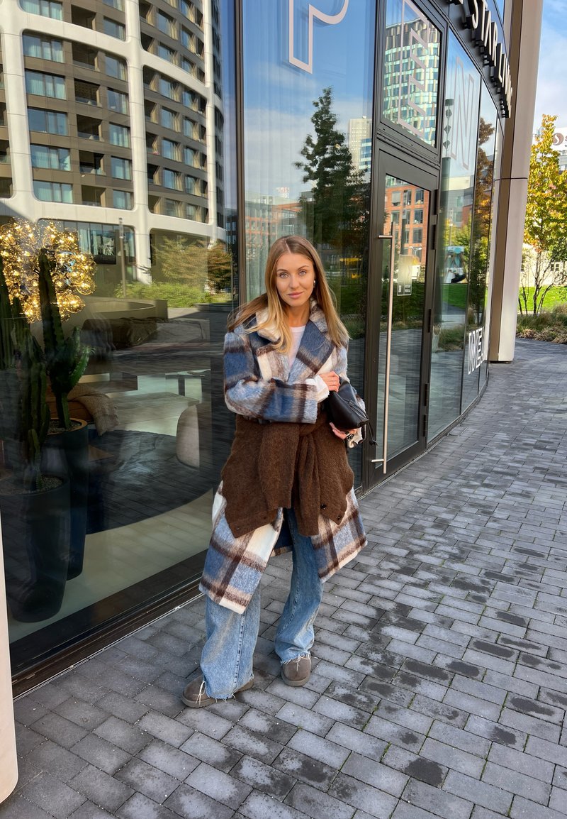 Καρό παλτό σε μπλε, λευκό και καφέ χρώμα με καφέ ζώνη, συνδυασμένο με φαρδιά μπλε τζιν και γκρι παπούτσια, όρθιο δίπλα σε μια γυάλινη βιτρίνα.