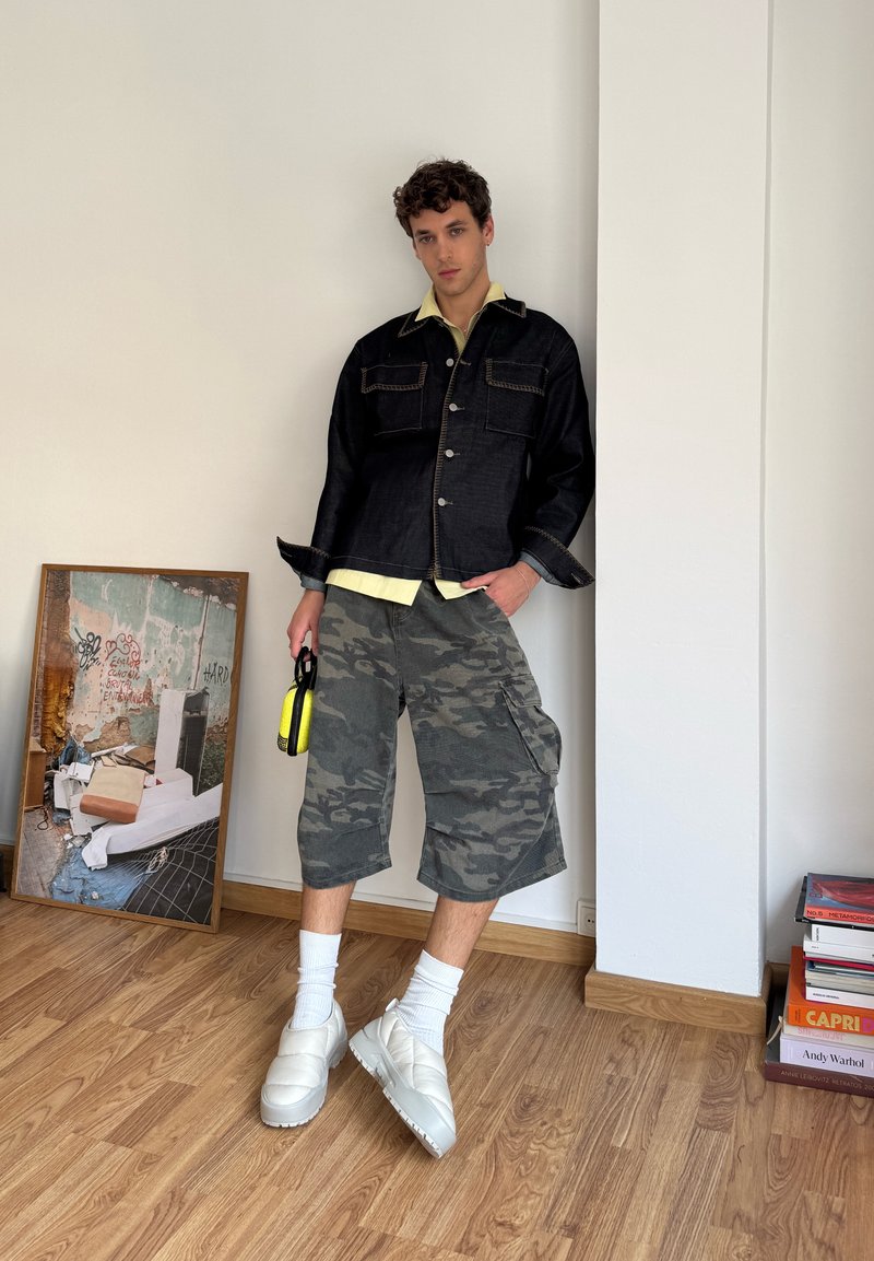 Giacca di jeans nera con due tasche sul petto, camicia gialla chiara, shorts cargo mimetici, scarpe bianche senza lacci, in mano una borsa gialla. Sfondo in legno.