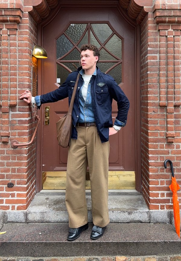 Jeune homme debout sur des marches devant une porte en bois, tenant la poignée de la porte d'une main et un parapluie orange à proximité à droite.