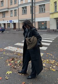 Casaco longo de pele preta sobre um conjunto preto, segurando uma bolsa castanho texturada. Asfalto molhado com folhas de outono caídas, ambiente urbano.