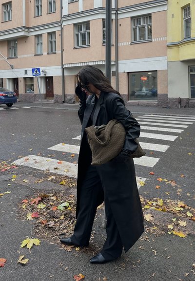 Abrigo largo de cuero negro sobre un conjunto negro, sosteniendo un bolso marrón texturizado. Asfalto mojado con hojas de otoño caídas, entorno urbano.