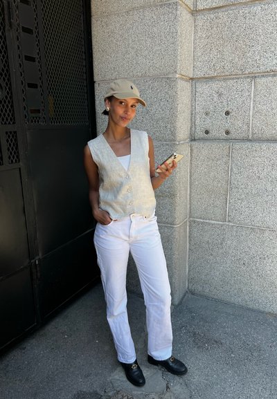 Chaleco de lino ligero con botones en el frente, camiseta blanca y pantalones blancos. Lleva una gorra beige y mocasines negros, sosteniendo un teléfono inteligente.