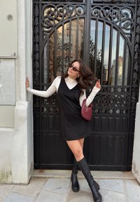 Rochie din suede negru cu un design în formă de V, suprapusă peste un pulover alb cu guler înalt. Cizme negre până la genunchi și o poșetă texturată burgundy. Poartă decorativă în fundal.