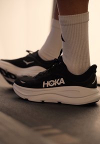 Chaussures de course noires avec semelles blanches épaisses. Caractéristiques : tige en maille, logo HOKA, et chaussettes blanches côtelées. Design minimaliste avec détails de marque.