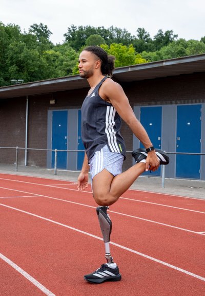 Atleta estirándose en una pista de atletismo, vistiendo una camiseta sin mangas negra y pantalones cortos claros, con una pierna protésica y zapatillas deportivas negras.