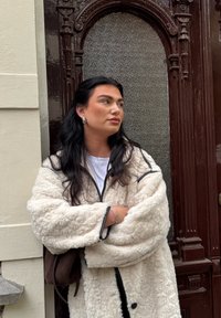 Casaco de lã branco e fofinho com acabamento preto, usado sobre uma blusa branca. A modelo tem cabelo longo e escuro e usa brincos de argola prateados. Porta de textura castanha ao fundo.