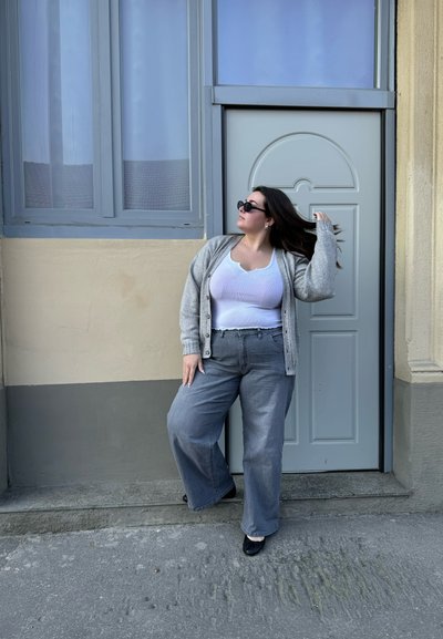 Cárdigan de punto gris sobre una blusa blanca, combinado con pantalones anchos grises y zapatos negros, en un entorno con una pared y una puerta de color claro.