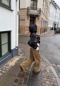 Άτομο που φοράει μαύρη κουκούλα και καφέ παντελόνι διασχίζει μια γωνία πεζόδρομου με πλακόστρωτο σε αστική περιοχή με κτίρια ευρωπαϊκού στιλ.