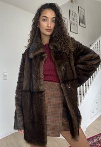 Abrigo de piel sintética marrón con cuello alto y grandes botones, llevado sobre un top burdeos de punto acanalado y una minifalda de cuadros. Tela texturizada, diseño de capas.