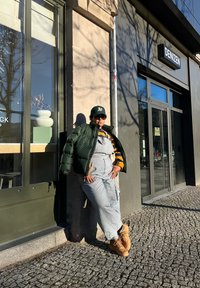 Πουκάμισο με ζακέτα σε σκούρο πράσινο, ριγέ κίτρινο και ναυτικό ζιβάγκο, γκρι φορμά, καφέ μπότες, γυαλιά ηλίου, βασισμένος σε έναν πέτρινο τοίχο.