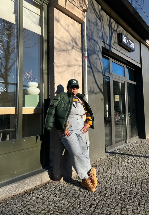 Puffer jacket in dark green, striped yellow and navy turtleneck, gray overalls, tan boots, sunglasses, leaning against a stone wall.