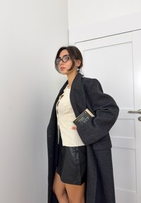 Dolga temno siva volnena plašč, svetlo rebrast pulover, črna usnjena mini krila. Drži knjigo; okrogla očala; minimalni dodatki; nevtralna ozadje.
