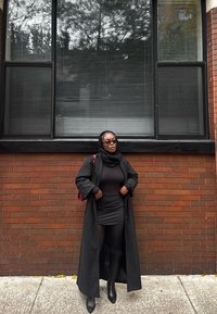 Cappotto lungo nero sopra un vestito nero, abbinato a collant neri e stivali al ginocchio. Accessoriato con uno zaino bordeaux. Muro di mattoni sullo sfondo.