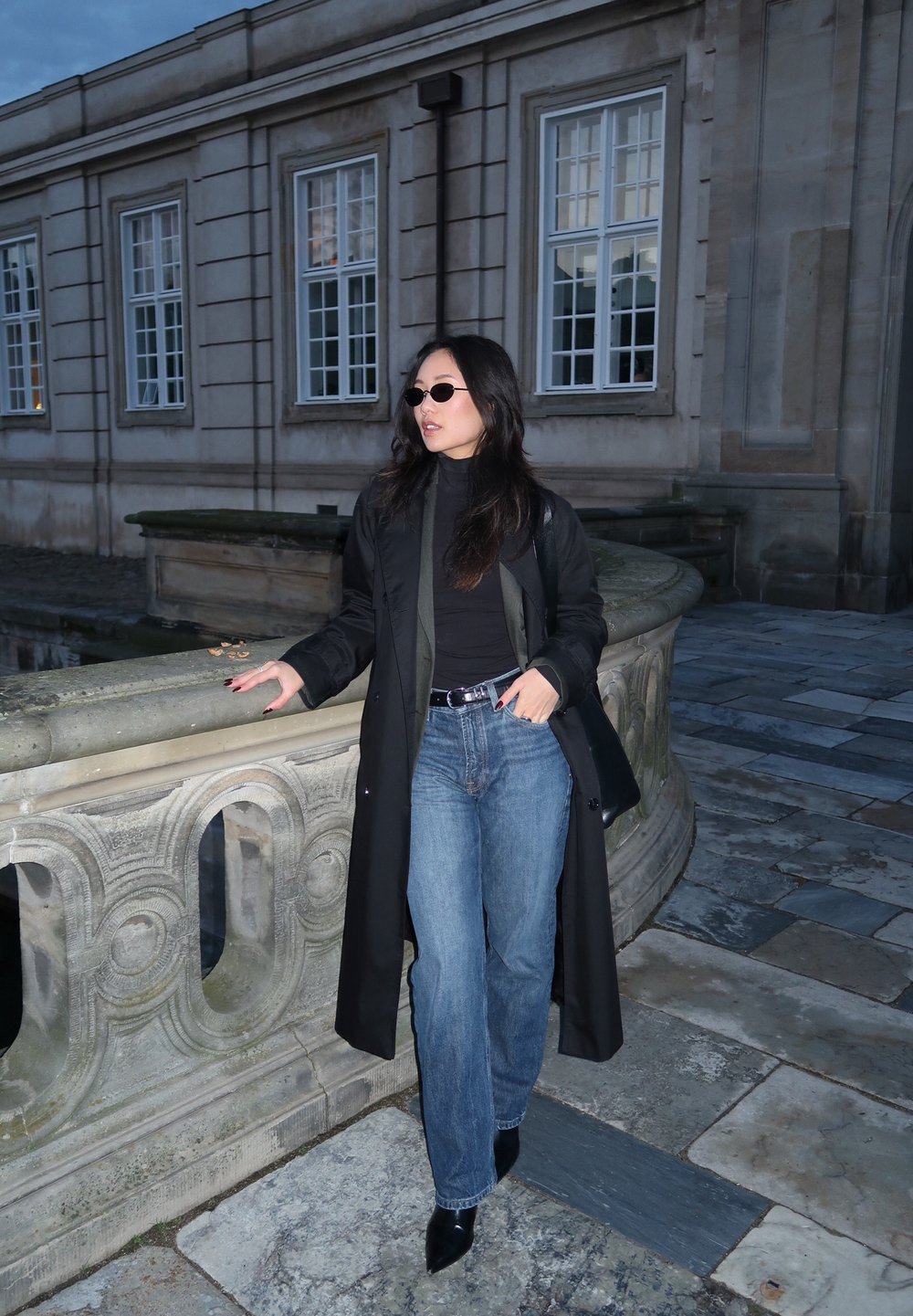 A black long coat worn over a black turtleneck, paired with blue straight-leg jeans and black pointed-toe boots. Accessories include round sunglasses.