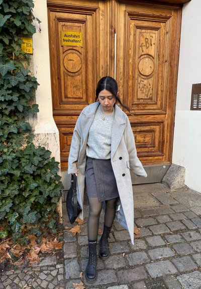 Abrigo de lana gris con solapa sobre un suéter claro. Falda negra, medias opacas y botas negras con cordones. El fondo incluye una puerta de madera y hiedra.