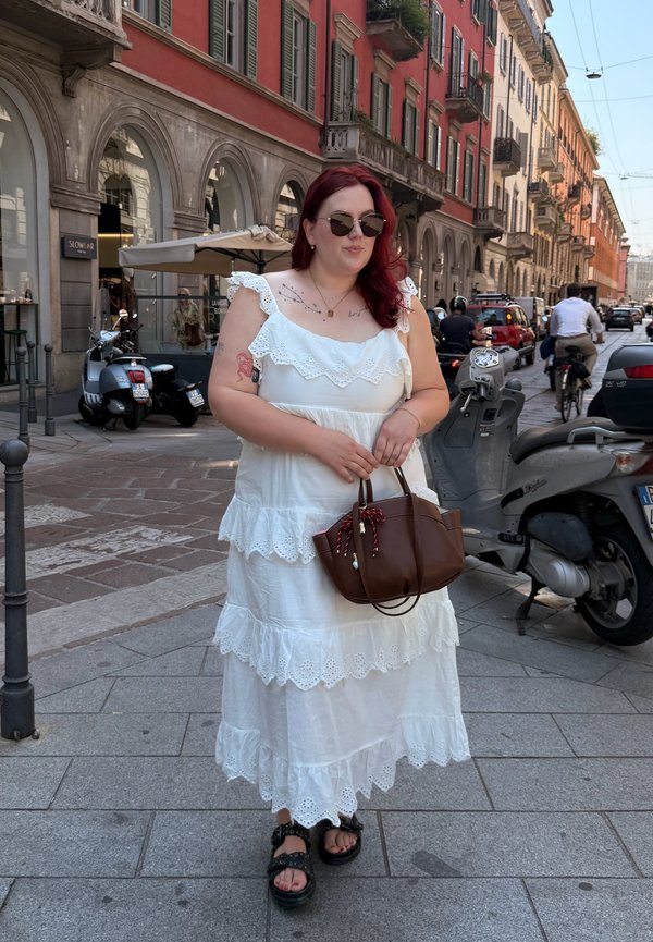Femme aux cheveux roux portant une robe blanche à volants et des lunettes de soleil, tenant un sac à main marron dans une rue de la ville avec des scooters et des cyclistes.