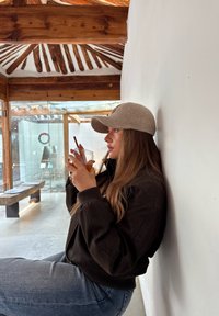 Femme portant une casquette grise et une veste sombre, assise contre un mur blanc, tenant un verre avec une paille, dans un café aux poutres en bois.