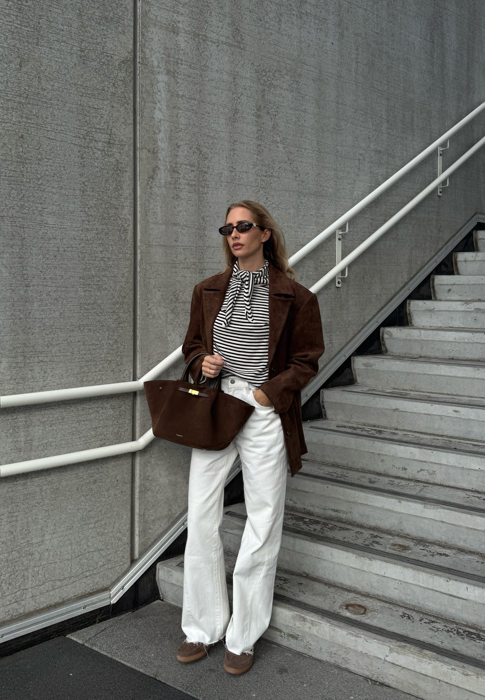 Brown suede coat over a black and white striped blouse, paired with white wide-leg trousers and a brown handbag. Accessories include black sunglasses.