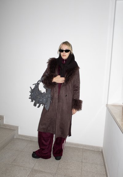 Mujer con abrigo marrón oscuro con ribete de piel, pantalones burdeos, pañuelo negro en la cabeza y gafas de sol, sosteniendo un bolso negro con textura, de pie en el interior.