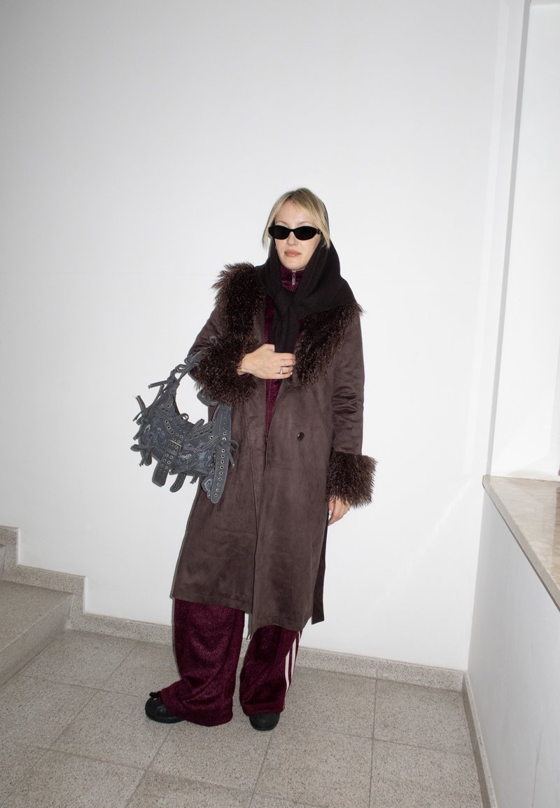 Femme portant un manteau marron foncé bordé de fourrure, un pantalon bordeaux, un foulard noir et des lunettes de soleil, tenant un sac à main texturé noir, debout à l'intérieur.