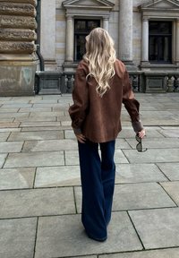 Cămașă oversized din suede maro cu mâneci lungi, asortată cu blugi evazați de culoare albastru închis. Textură netedă și croială lejeră, stând pe un trotuar de piatră.