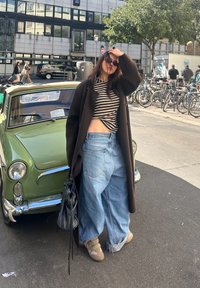 Carro vintage verde ao fundo; pessoa vestindo um casaco longo preto, blusa às riscas, jeans de perna larga azul clara e calçado bege, segurando uma bolsa preta.