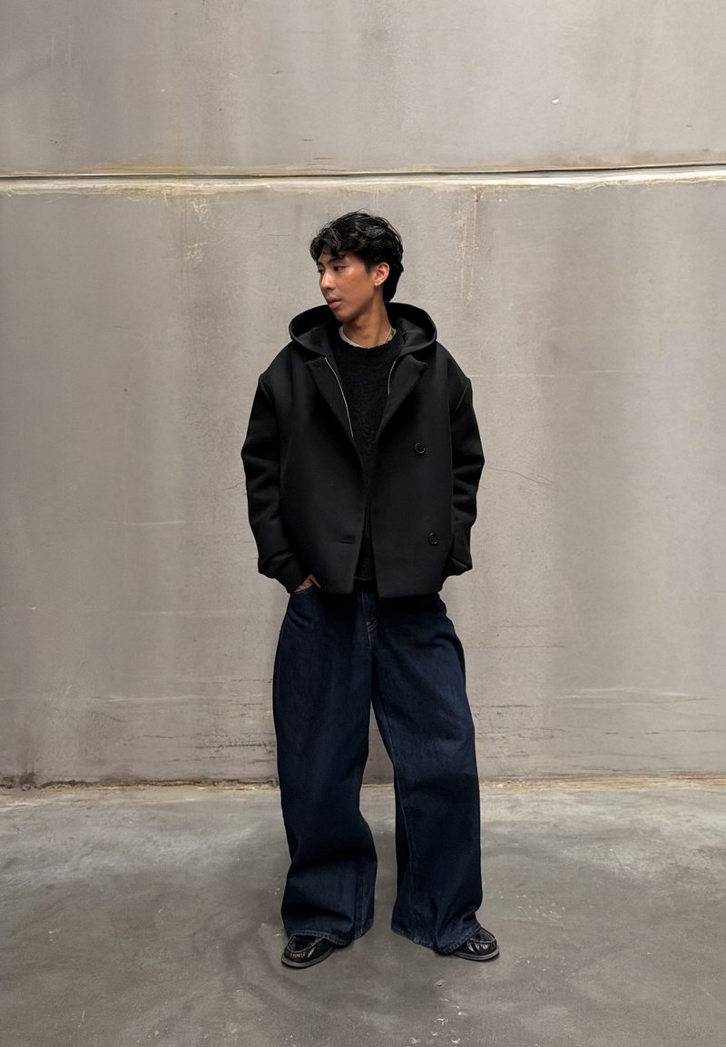 Jeune homme portant une veste à capuche noire et un jean large foncé, debout les mains dans les poches, regardant sur le côté contre un mur uni.