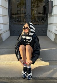 Fekete bőrdzseki, csíkos fekete-fehér kötött kapucnis pulóver, fekete sneaker fehér fűzővel, kőlépcsőn ülve.