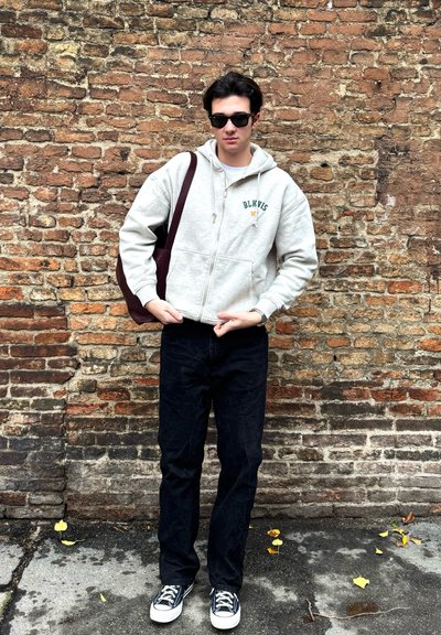 Sudadera gris con cremallera y texto verde, jeans oscuros y zapatillas negras, sosteniendo una bolsa de lona marrón contra un fondo de pared de ladrillo.