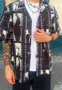 Άνδρας που φοράει πουκάμισο με εκτύπωση ταινίας ασπρόμαυρης, από πάνω σε λευκό μπλουζάκι, πολύχρωμα κολιέ από χάντρες και τατουάζ στους πήχεις.