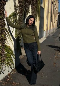 Olivgrüner gerippter Cardigan mit Kunstfellkragen, kombiniert mit dunkelblauen, ausgestellten Jeans. Schwarze Handtasche und Blätter im Hintergrund.