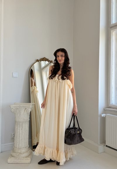 Mujer con cabello largo y oscuro con un vestido fluido color crema, sosteniendo un bolso negro, parada junto a un pedestal blanco decorativo y un espejo alto.