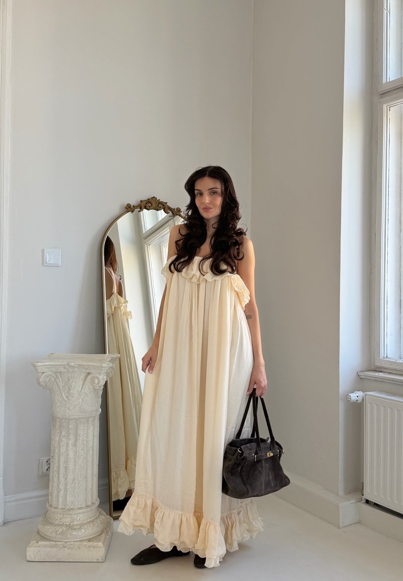 Donna con lunghi capelli scuri, che indossa un abito crema vaporoso e tiene in mano una borsa nera, in piedi accanto a un piedistallo bianco decorativo e a uno specchio alto.