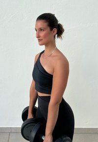 Woman in black workout gear standing against a white wall, holding dumbbells and facing sideways with her hair tied back.
