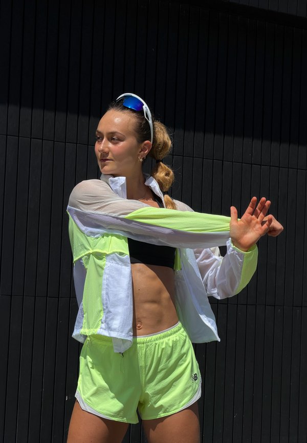 Femme en tenue de sport jaune néon étirant ses bras devant un mur noir, portant des lunettes de soleil sur la tête et une queue de cheval.