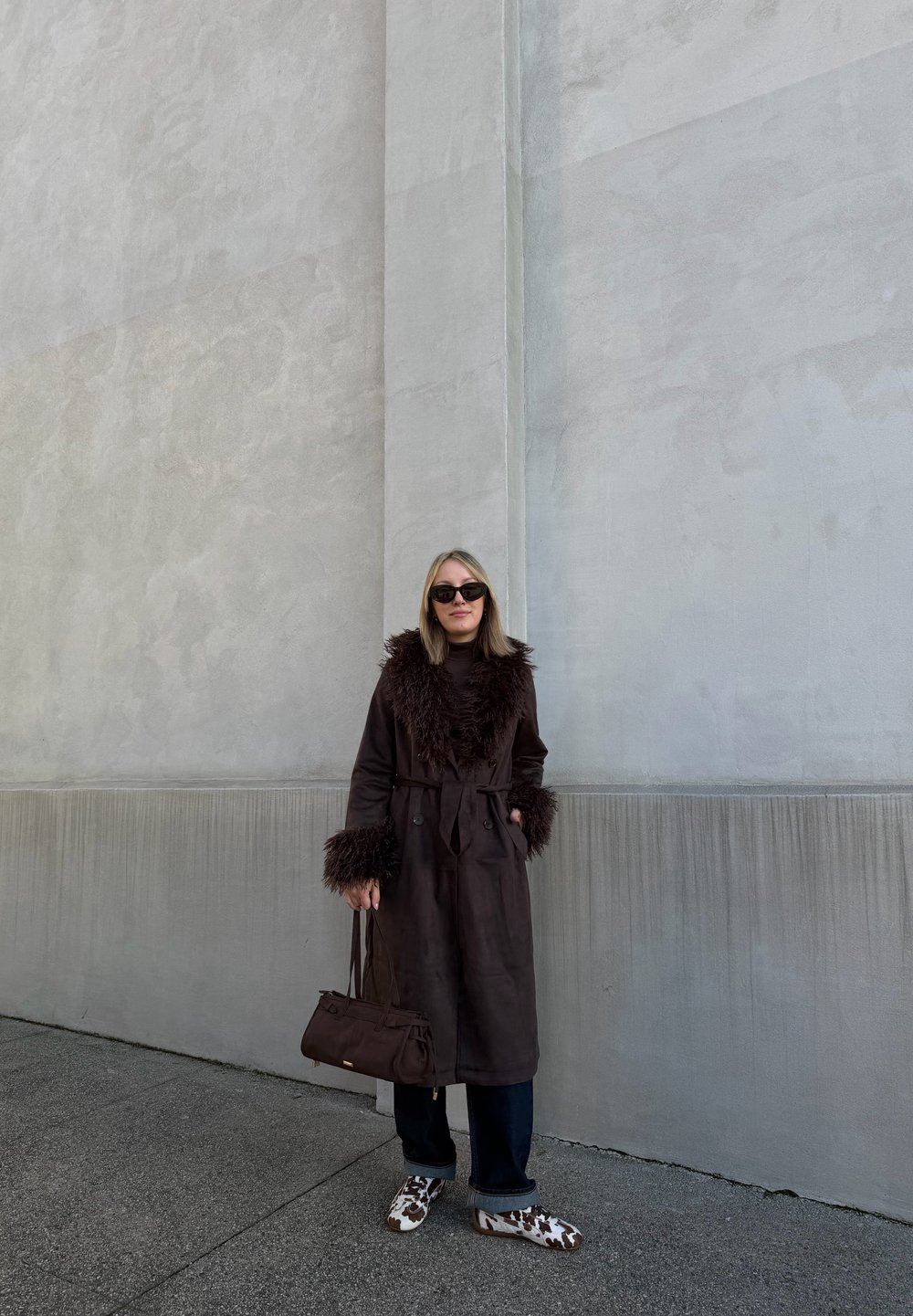 Barna trench kabát bolyhos gallérral és mandzsettákkal, fekete farmer, és barna kézitáska. A lábbeli fehér és barna pöttyös mintázattal rendelkezik.