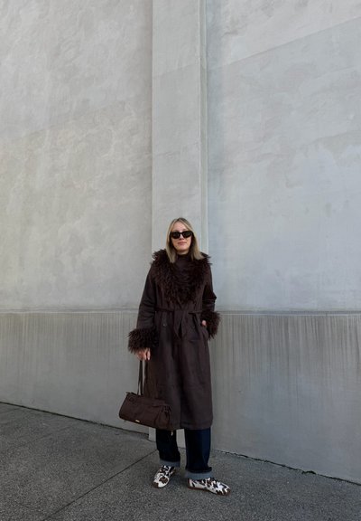 Trench de color marrón con un cuello y puños de piel, jeans negros y un bolso marrón. Footwear presenta un patrón moteado en blanco y marrón.