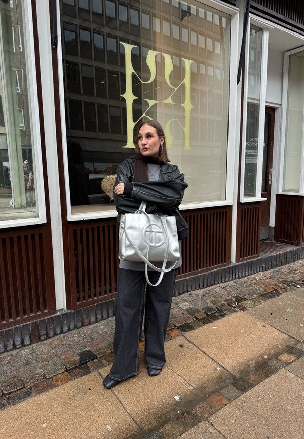 Femme portant une veste en cuir noire et un jean large se tient sur un trottoir en pierre devant une vitrine avec de grandes lettres jaunes.