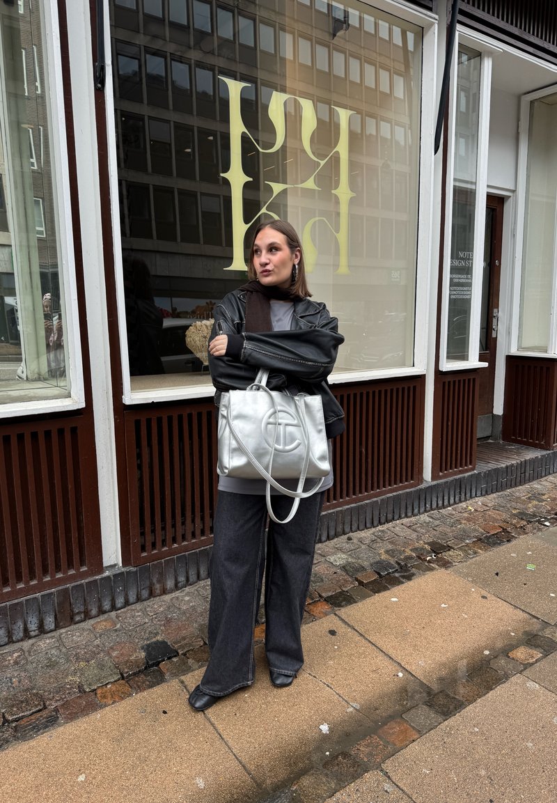 Woman wearing black leather jacket and wide-leg jeans stands on stone sidewalk in front of store window with large yellow letters.