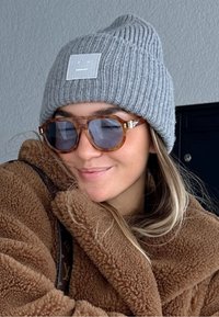 Pelēks adīta cepure ar kvadrātveida uzšuvi, brūna flīsa jaka un bruņurupuču stila saulesbrilles ar zilām lēcām, demonstrējot slāņainu ziemas izskatu.