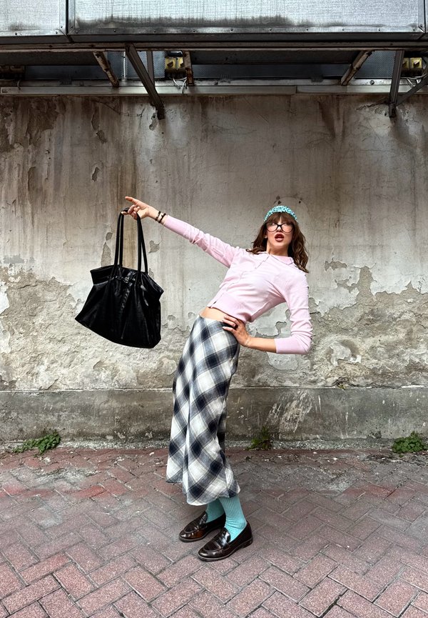 Femme portant un haut rose, une jupe à carreaux, des chaussettes bleues et des lunettes, dansant et tenant un grand sac noir devant un mur en béton usé.