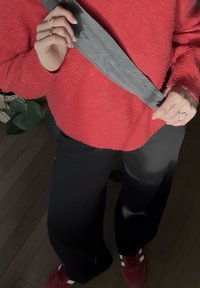 Piros bolyhos pulóver, fekete bőszárú nadrág, szürke öv és piros, fehér csíkos tornacipő. A háttérben fa padló és növénylevelek láthatók.