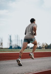 Un athlète en chemise marron clair et short vert court sur une piste rouge. Ses baskets sont principalement blanches avec des accents noirs et gris clair.