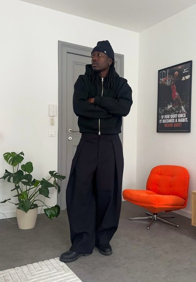 Una persona está de pie vistiendo una sudadera negra con cremallera y pantalones anchos negros. Lleva un gorro negro y zapatos negros resistentes, en una habitación con plantas.