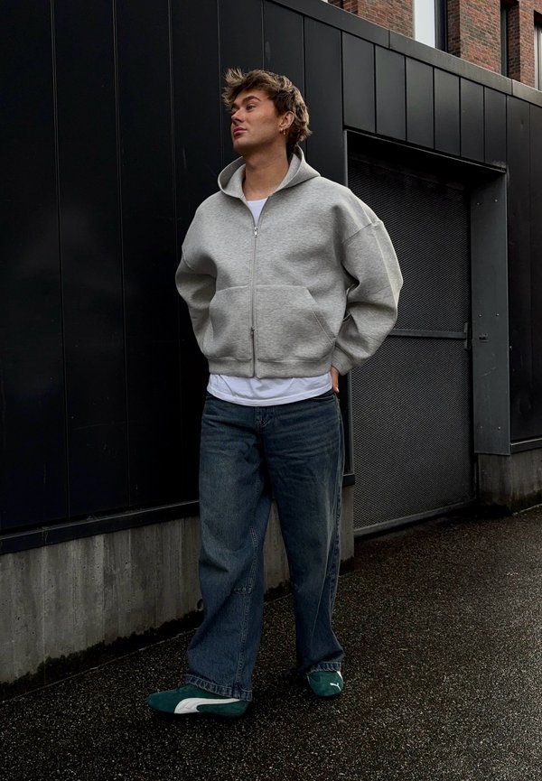 Casaco cinza com fecho, T-shirt branca, jeans azuis largos e ténis verdes. Conjunto casual com ajuste relaxado e textura suave. Fundo urbano.