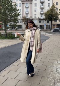 Trench coat beige con grandi bottoni, sciarpa a quadri beige e marrone, jeans scuri e ballerine nere, mentre tiene una tazza in un contesto urbano.