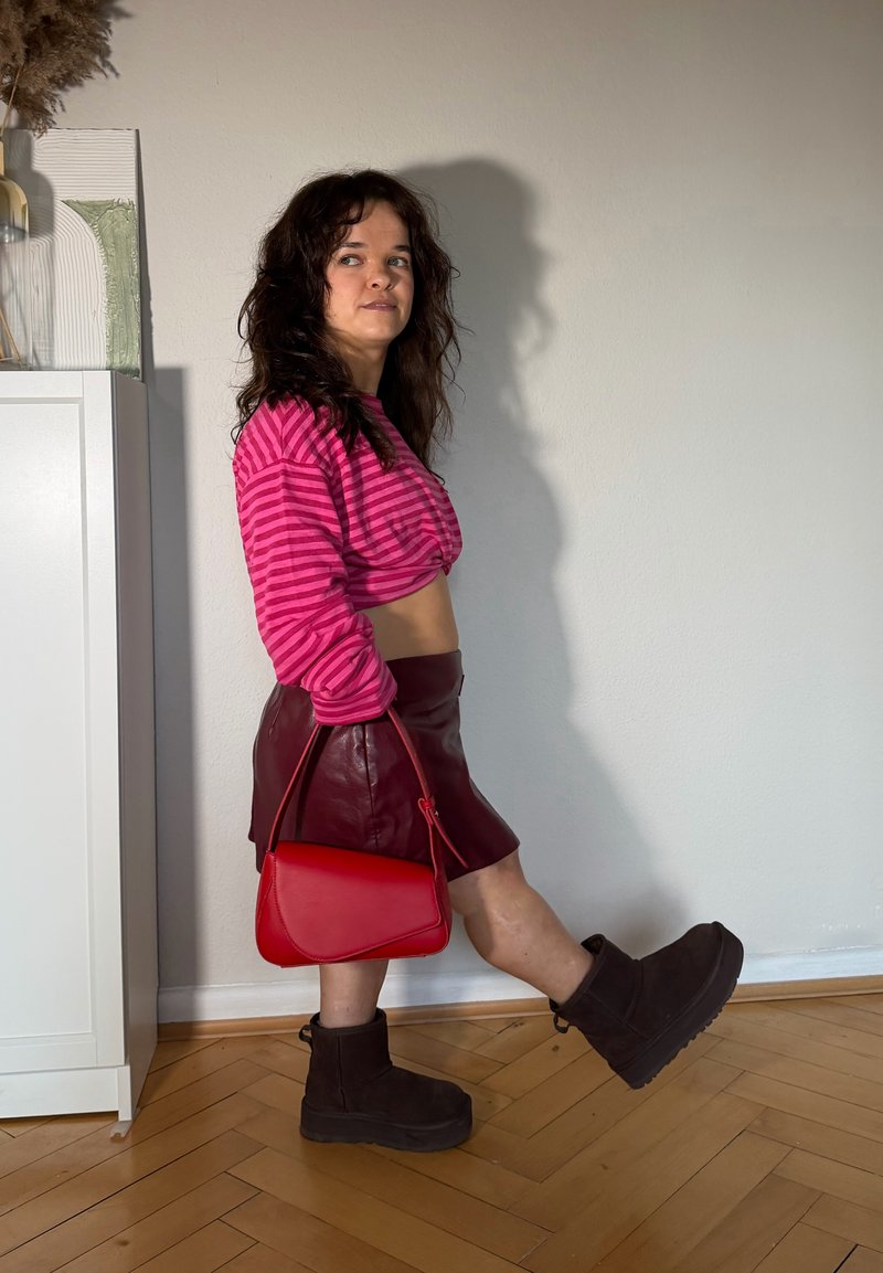 Jeune femme en haut court à rayures roses, jupe bordeaux, tenant une pochette rouge, portant des bottes marron foncé, debout sur un sol en bois à l'intérieur.