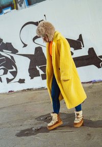 Casaco de polar amarelo com collar grande, top laranja, jeans azuis e botas de inverno castanhas com detalhes em pelo, contra um muro coberto de graffiti.